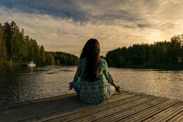 Frau meditiert am Ufer eines ruhigen Sees bei Sonnenuntergang.